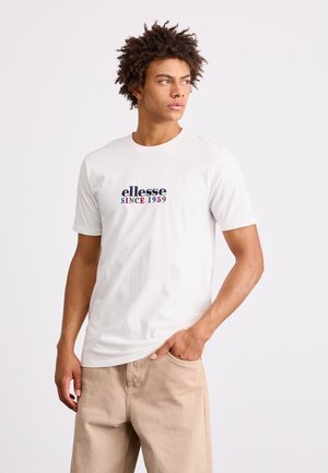 T-shirt bianca in cotone con maniche corte, caratterizzata dal logo ricamato "ellesse" in blu e rosso. Indossata con pantaloni beige, dalla texture liscia.