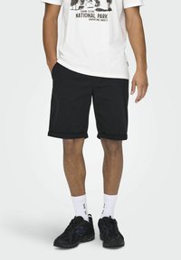 Pantalones cortos negros con dobladillos enrollados, hechos de una tela sólida. Se llevan con una camiseta gráfica blanca, zapatos deportivos negros y calcetines blancos.