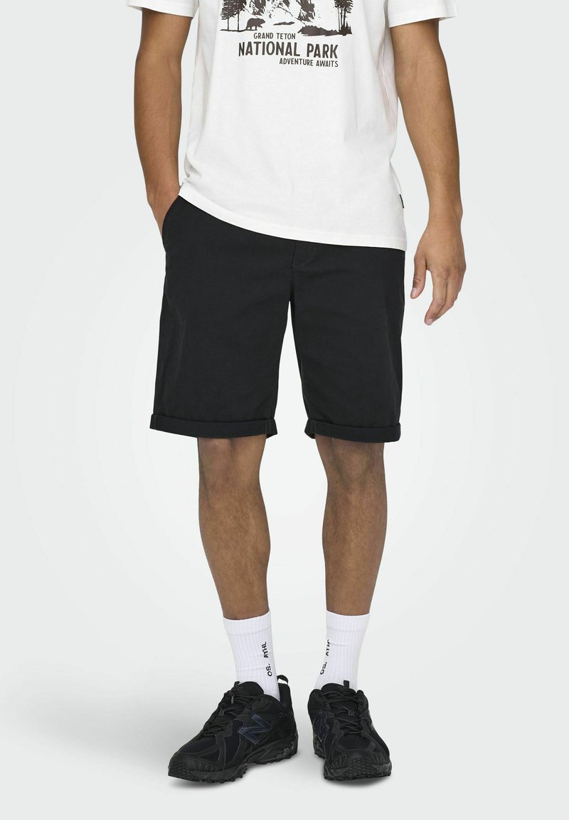 Pantalones cortos negros con dobladillos enrollados, hechos de una tela sólida. Se llevan con una camiseta gráfica blanca, zapatos deportivos negros y calcetines blancos.