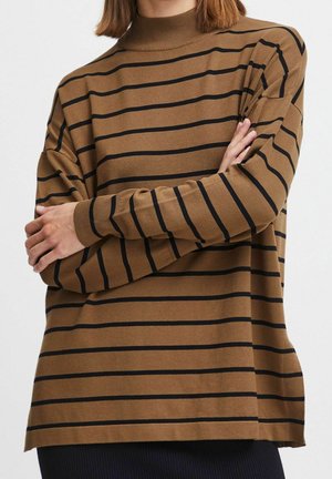 Person trägt einen braunen Pullover mit langen Ärmeln und schwarzen horizontalen Streifen, die Arme verschränkt, das Bild ist am Hals und an den Hüften beschnitten.