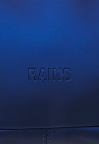 Niebieski wodoodporny materiał o gładkiej fakturze z wytłoczonym logo "RAINS" na powierzchni, podkreślający prostotę i nowoczesny design.