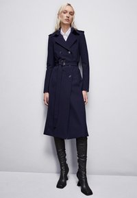 Trench coat blu navy con bottoni doppi, cintura e spalline, abbinato a stivali neri al ginocchio. Tessuto liscio.