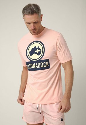 Hombre con camiseta rosa claro con logo de Altonadock en azul oscuro y pantalones cortos a juego sobre fondo liso.