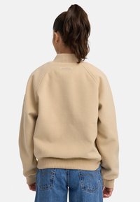 Sweatshirt beige avec col et poignets côtelés, présentant une étiquette visible au dos. Porté avec un jean bleu, montrant une coupe décontractée et une texture.