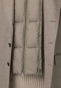 Cappotto esterno beige con tessuto texture e bottoni neri, aperto per rivelare uno strato interno imbottito di un grigio chiaro dal finish liscio.