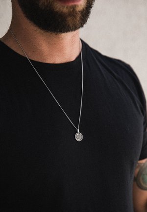 Silver necklace with a round pendant featuring a detailed design, worn over a black t-shirt. Chain is fine and textured.