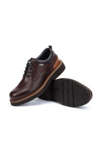 Chaussures en cuir marron avec une finition lisse, bout rond et un design à lacets. Elles présentent un talon marron contrastant et une semelle en caoutchouc texturée.