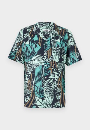 Mann trägt ein kurzärmeliges Hemd mit tropischem Muster, marineblaue strukturierte Shorts und braune Ledersandalen, steht mit den Händen in den Taschen.