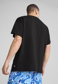 Sort bomulds T-shirt med afslappet pasform, korte ærmer og rund hals. Har et mærkelabel ved kanten; båret med blå mønstrede shorts.
