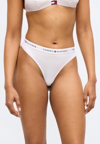 Balts tangas apakšveļas briefs no mīksta auduma ar zīmola elastīgo jostu, uz kuras ir uzraksts "TOMMY HILFIGER" treknā tekstā.
