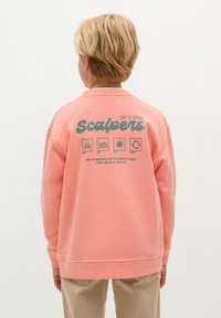 Blond dziecko stoi tyłem, ubrany w koralowy sweatshirt z logo "Scalpers" oraz ikonami czaszki, zabawy, słońca i powtórzenia na plecach, w połączeniu z beżowymi spodniami.