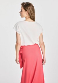 Biała bluzka z krótkim rękawem zestawiona z koralową spódnicą w stylu wrap, posiadającą boczne wiązanie i miękką fakturę, widziana od tyłu.