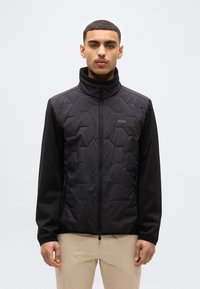 Νέος άνδρας με κοντά μαλλιά, φορώντας μαύρο κολεγιό με ψηλό γιακά και μπεζ παντελόνι, στέκεται μπροστά σε απλό φόντο.