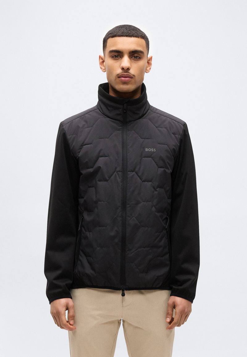 Νέος άνδρας με κοντά μαλλιά, φορώντας μαύρο κολεγιό με ψηλό γιακά και μπεζ παντελόν ι, στέκεται μπροστά σε απλό φόντο.