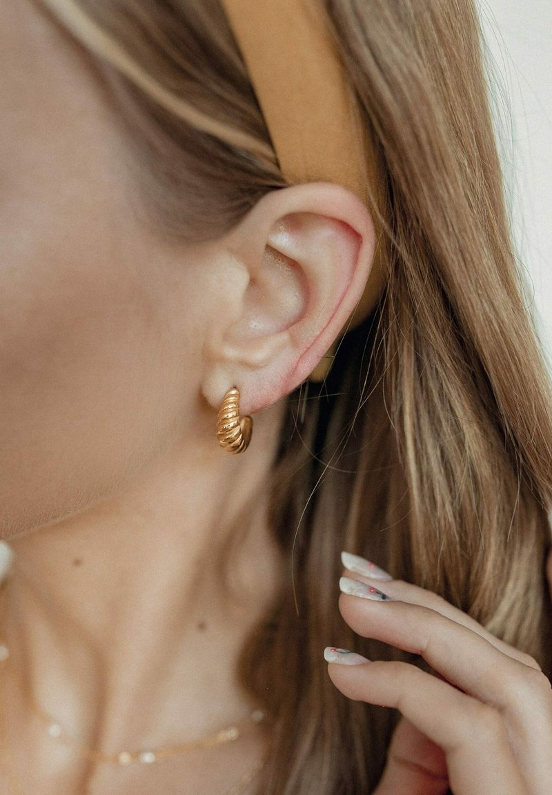 Boucle d'oreille en spirale dorée sur l'oreille d'un modèle ; texture métallique lisse ; le gros plan révèle des détails de crêtes ; cheveux clairs et main visibles.
