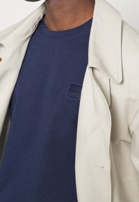 Maglietta di cotone blu navy con un'etichetta quadrata con la scritta "BOSS." Giacca esterna beige con dettagli di cuciture visibili e collo a revers.