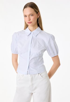 Młoda kobieta w białej dopasowanej bluzce z krótkim rękawem w niebieskie paski i białych, wysokich spodniach, stoi z rękami za plecami.