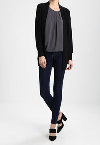 Schwarzer offener Cardigan, graue Bluse mit Falten, dunkle schmale Jeans und schwarze High Heels mit Knöchelriemen. Minimalistisches Design.