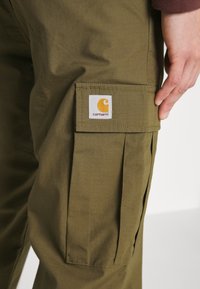 Olivgrüne Cargo-Hosen aus strukturiertem Stoff. Verfügt über eine große Cargotasche, Falten und ein sichtbares Carhartt-Logopatch.