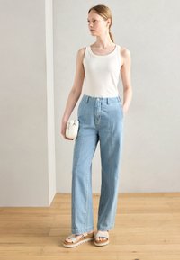 Cream tank top, high-waisted light blue wide-leg jeans, and tan platform sandals. Model holds a small white bag. Wood floor and neutral background.