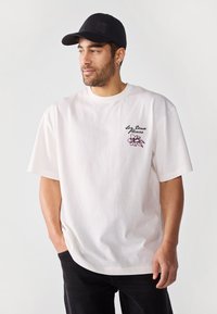 White cotton t-shirt with short sleeves featuring a purple floral embroidery and text design on the left chest. Paired with a black cap.