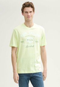 Lysegrøn bomulds T-shirt med korte ærmer, prydet med en hvid trykt logo tekst og detaljer. Båret med blå jeans.