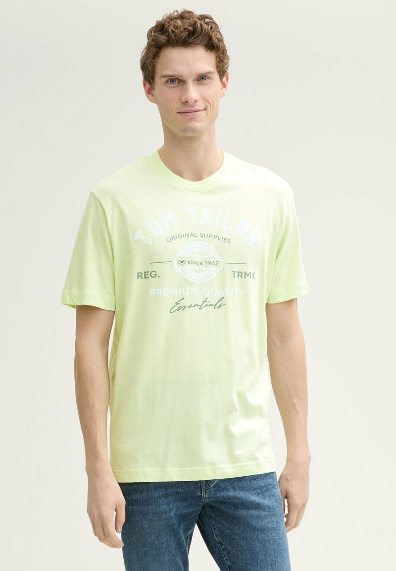 Lysegrøn bomulds T-shirt med korte ærmer, prydet med en hvid trykt logo tekst og detaljer. Båret med blå jeans.