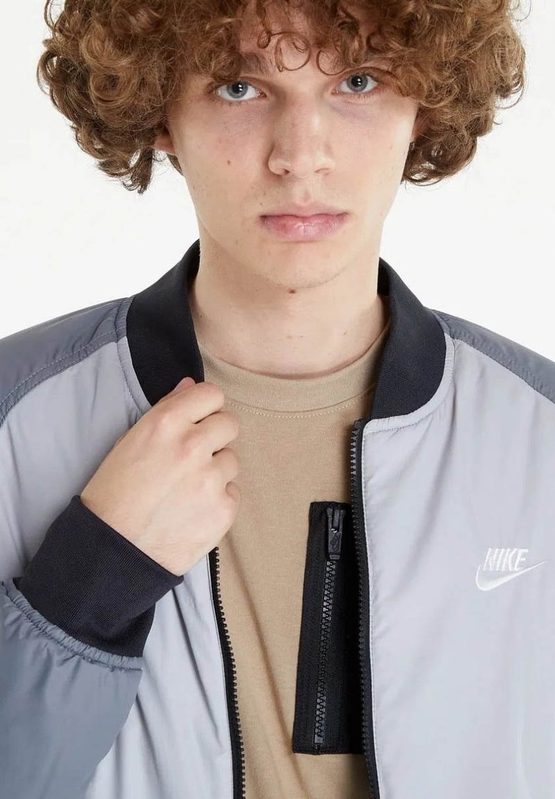 Giovane con capelli ricci che indossa una giacca Nike grigia sopra una maglietta beige, mentre tiene il colletto della giacca con una mano.