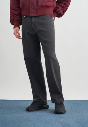 Άτομο που φορά φαρδιά μαύρα τζιν, μαύρα αθλητικά παπούτσια και κόκκινο μπουφάν, στέκεται σε μπλε χαλί.