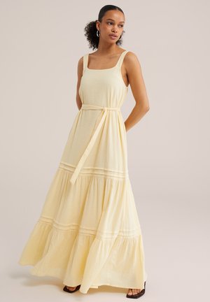 Femme portant une robe sans manches, jaune pâle, longueur cheville, avec un décolleté carré et une ceinture nouée à la taille, debout devant un fond uni.