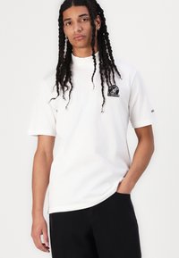 Tricou alb cu mânecă scurtă, având un guler rotund și o imprimare grafică neagră pe partea stângă a pieptului. Materialul pare moale și neted.