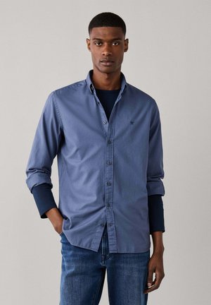 Mann trägt ein blaues Hemd mit Knöpfen über einem dunklen Langarmshirt und blauen Jeans, steht mit einer Hand in der Tasche vor einem schlichten Hintergrund.