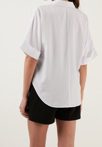 Weißen Kurzarmbluse mit abgerundetem Saum und glatter Textur. Mit einem Rückennaht-Detail, kombiniert mit schwarzen Shorts.