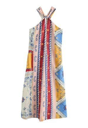 Nő, színes foltvarrott pántos maxi ruhában, fekete kézitáskát tartva, napsütötte, mintás csempézett udvaron állva.