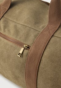 Sac de voyage en toile vert olive avec une poignée tissée marron. Doté d'une fermeture éclair bronze avec un curseur et de détails de design cousus.