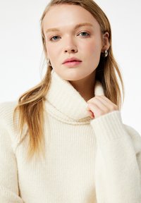 Suéter de cuello alto color marfil con textura acanalada, cuello grueso y mangas largas, que presenta una tela suave y un corte relajado.