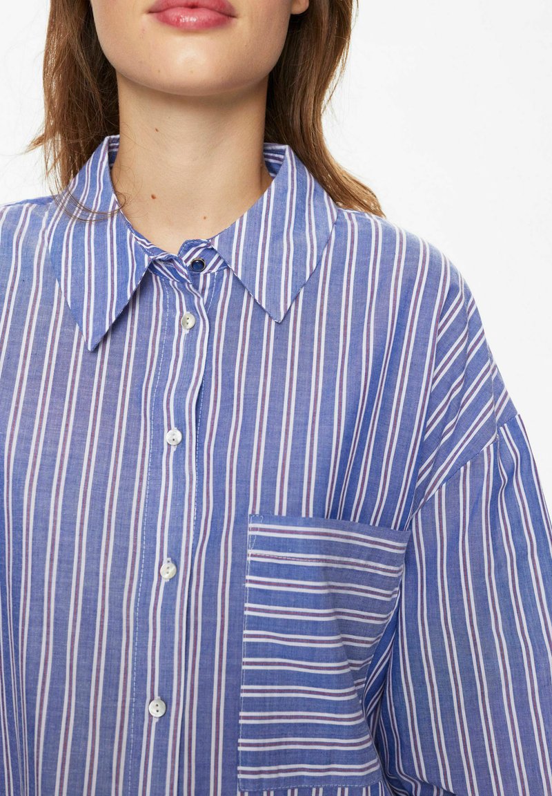 Camicia a strisce blu realizzata in tessuto leggero, con colletto a punta, tasca frontale e righe orizzontali e verticali di larghezze variabili.