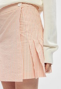 Lichtroze rok met verticale witte strepen, voorzien van een knoopsluiting en gestructureerde plooien. Het materiaal lijkt gestructureerd en gekreukt te zijn.