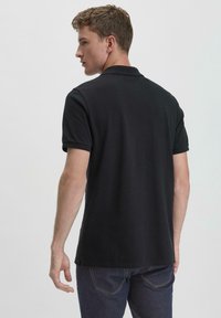 Black polo shirt with a classic design, short sleeves, and a ribbed collar, paired with dark jeans, viewed from the back.