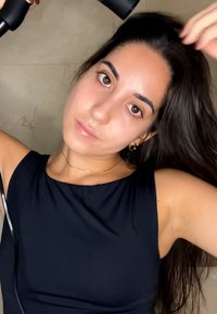 Vrouw met lang donker haar draagt een zwart mouwloos topje en gebruikt een föhn om haar haar te drogen tegen een beige betegelde achtergrond.