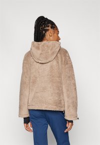 Beige Kunstfelljacke mit Kapuze, im entspannten Schnitt, mit strukturierter Oberfläche und gerippten Bündchen, über blauen Hosen getragen.