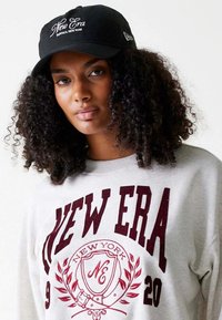 Cappellino nero con "New Era" ricamato in bianco, indossato con una felpa grigia chiara caratterizzata da scritte bordeaux e un design di stemma decorativo.
