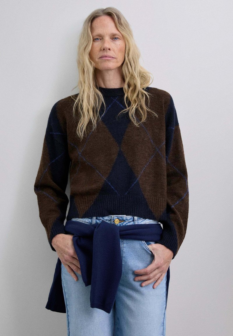 Pull en motif losange marron et marine avec un col et des poignets côtelés, associé à un jean taille haute bleu clair et un châle tricoté marine.