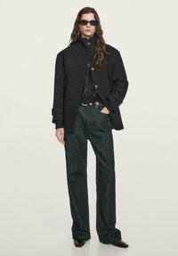 Cappotto nero oversize con colletto alto, maglione grigio a maglia sotto, pantaloni larghi verde scuro e scarpe nere eleganti. Occhiali da sole alla moda completano il look.