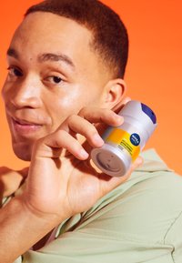 Antitraspirante NIVEA Men in un contenitore cilindrico grigio con un tappo blu e un'etichetta gialla, tenuto da una persona su uno sfondo arancione.