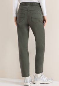 Personne portant un pantalon vert olive avec des poches arrière, des chaussettes blanches, des baskets argentées et un haut blanc à manches longues, debout contre un mur beige.