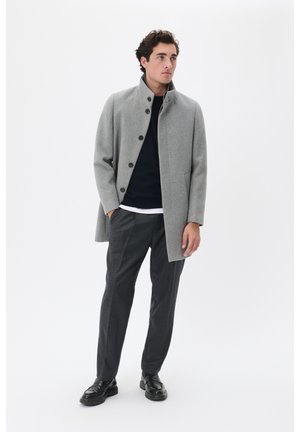 Grau melierter Wollmantel mit hohem Kragen, vorderen Knöpfen und seitlichen Taschen, getragen über einem dunkelblauen Pullover und grauen Hosen, kombiniert mit schwarzen Schuhen.