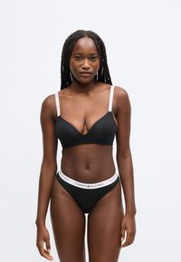 Sort trådløs bh med V-udskæring og justerbare hvide stropper med mærke-logo. Matchende sort g-streng med hvidt logo-bånd.