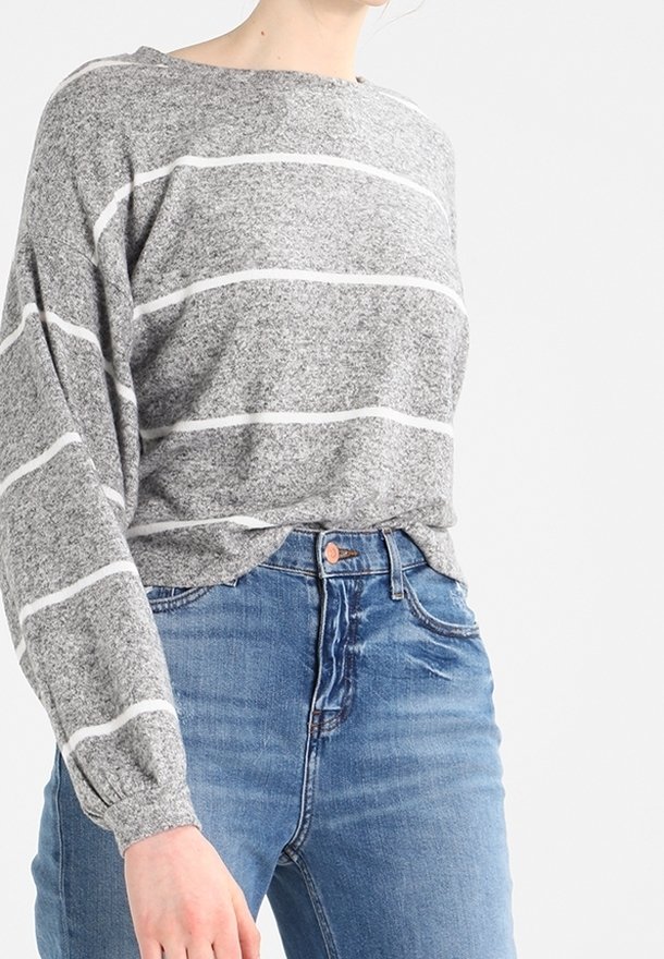Personne portant un pull gris à rayures horizontales blanches et un jean bleu taille haute, debout devant un fond uni.