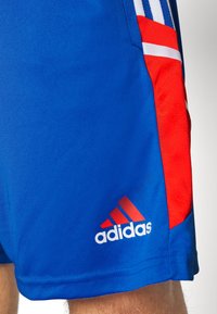 Modré sportovní šortky Adidas s červenými akcenty, které nosí osoba, s logem Adidas poblíž spodního okraje.